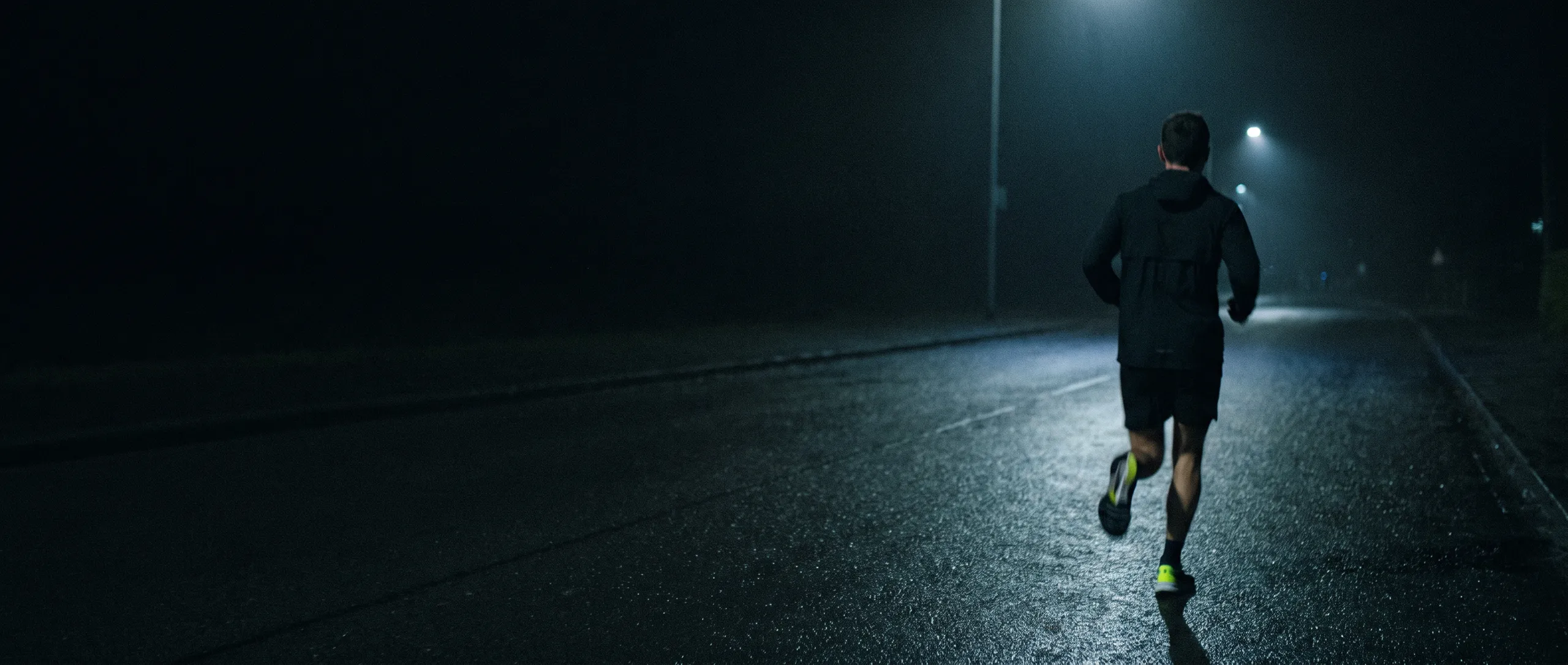 A runner on wet asphalt at night, seen from behind under a single streetlight.